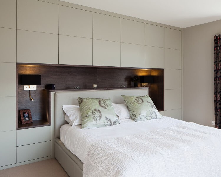 A modern bedroom with a light grey bed and matching fitted furniture. The bed has white bedding and two green pillows with floral patterns. The headboard, part of the fitted cabinetry, creates a recessed shelf with lighting on each side. A framed photo sits on the left shelf, enhancing this elegant fitted bedroom design.