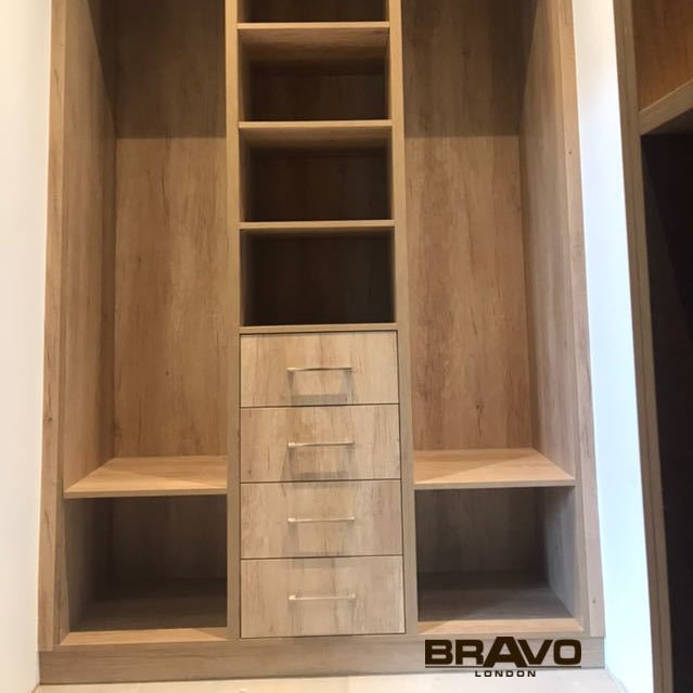 A custom-built wooden wardrobe featuring a combination of shelves and drawers, with a lighter wood finish, installed in a room corner, displaying the logo "Bravo London" on the bottom right. Ideal for fitted bedrooms.