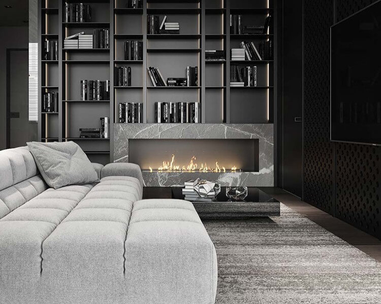 A modern living room featuring a large gray sectional sofa with a book and glasses on a sleek coffee table in front. Behind the sofa is a minimalist bookshelf filled with books, and a linear fireplace integrated into a gray marble structure below the shelves, complementing the fitted furniture design.