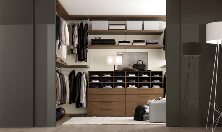 Modern, organized walk-in closet with hanging clothes, neatly folded items on shelves, and a multi-compartment drawer unit. A cushioned bench sits on a white rug, a standing lamp is positioned on the right, and storage boxes are on the top shelves—all showcasing exquisite fitted furniture.
