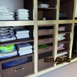 A neatly organized wardrobe featuring shelves filled with neatly folded towels, shirts, and other linens. Several compartments with closed drawers enhance the tidy look, complemented by a bespoke furniture design that maximizes space utilization.