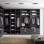 A modern, stylish walk-in wardrobe with neatly arranged clothes, shoes, and accessories. Features dark wood shelving, drawers, and a plush beige carpet.