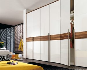 A modern bedroom featuring a large white wardrobe with horizontal wooden accents and sliding door wardrobes, a yellow bedspread, and scattered shoes and clothing items. Bright, stylish interior with Venetian blinds.