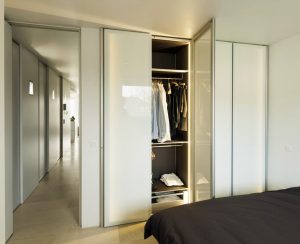A modern bedroom with a large, open wardrobe showing neatly arranged clothes and multiple shelves, lit by soft lighting, with a view into a corridor featuring bespoke furniture.