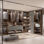 A spacious, modern walk-in wardrobe featuring wooden shelves filled with neatly organized clothes, glass sliding doors, and a center ottoman on a rug.