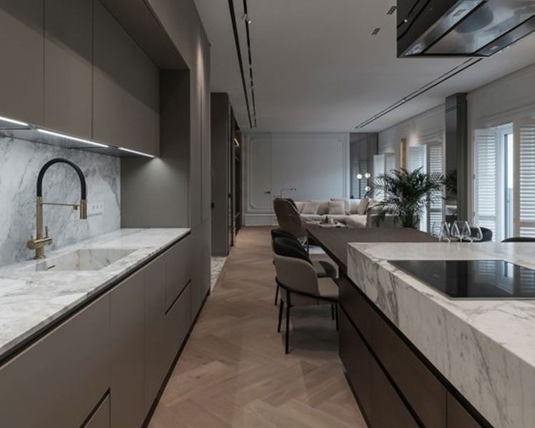 A modern kitchen with sleek, dark cabinetry and marble countertops. A built-in sink with a high-end faucet is on the left, and a marble island with a stovetop is on the right. The space extends to a stylish living area featuring fitted furniture, large windows, and light-colored pieces.