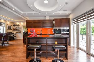 A modern kitchen with dark wood cabinets, an orange backsplash, and a central island with bespoke bar stools. It features recessed lighting and large windows letting in natural light.