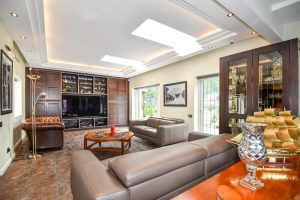 A luxurious living room featuring a brown leather sofa set, a bespoke glass coffee table, dark wood shelves, a large TV, and elegant ceiling recessed lights, with expansive windows allowing ample natural light.