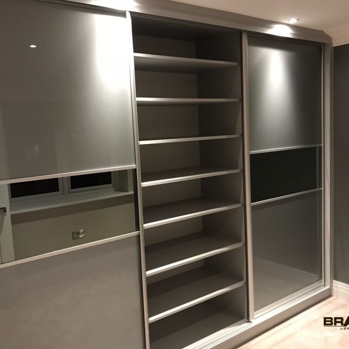 Modern fitted wardrobe with sliding frosted glass doors and multiple shelving units, installed in a well-lit room with grey walls.