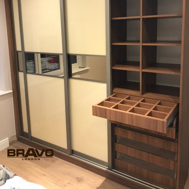 A modern wooden wardrobe with sliding doors is partly open, revealing neatly organized shelves and compartments. A pull-out drawer with multiple sections is extended. A bronzed mirror accents the interior, adding a touch of elegance. A logo in the bottom left corner reads "BRAVO LONDON." The flooring is light-colored wood.