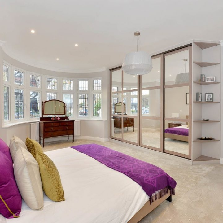 A spacious, modern bedroom featuring a king-sized bed with purple bedding, large windows, fitted wardrobes with sliding doors, and stylish hanging lamps, set in a room with soft gray walls and hardwood floors.