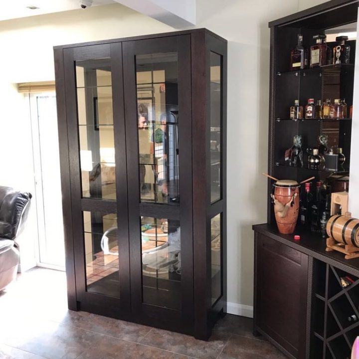 A tall, dark wooden display cabinet with glass doors stands between a liquor shelf and a leather chair in a well-lit room with bespoke furniture and tiled flooring.