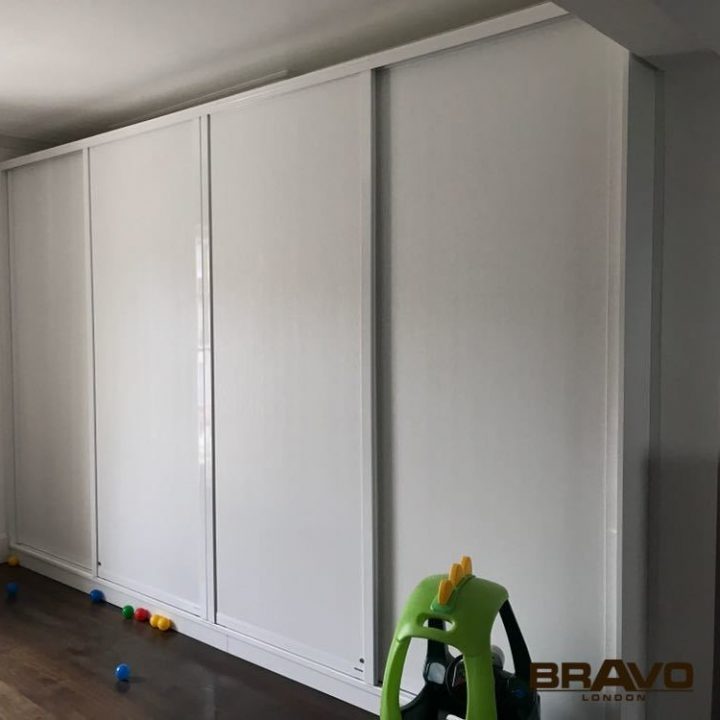 A room featuring large white hinged door wardrobes in a clean, modern design, with a wooden floor. A green and yellow toy car and scattered colorful balls are visible on the floor.