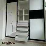 Modern white and black fitted wardrobe with open drawers and shelves in a bright, minimalistic room with a visible bathroom in the background.