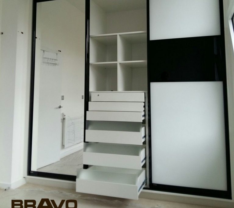 Modern white and black fitted wardrobe with open drawers and shelves in a bright, minimalistic room with a visible bathroom in the background.