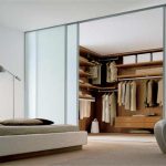 A modern bedroom with a large sliding door wardrobe open to reveal neatly organized clothes. A stylish chair and a low bed with beige linens add to the minimalistic decor.