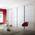 Modern living room with a bold red wall, large white sliding door panels concealing fitted wardrobes, a red modern chair, transparent glass table, and decorative vases on a neutral rug.