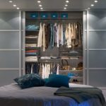 A modern and tidy bedroom featuring a large, organized closet with clothes and accessories visibly arranged behind hinged door wardrobes, complemented by soft lighting and a neatly made bed in the foreground.