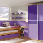 A modern bedroom with a purple and white theme, featuring sliding door wardrobes, a bed with storage underneath, floating shelves, and desk area by the window.