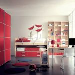 A modern bedroom featuring a red sliding door wardrobe, a white loft bed with a desk and computer underneath, a shelving unit filled with books and assorted items, and a red rolling suitcase.