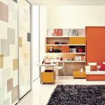 A modern living room with orange and white decor featuring a geometric wall design, a built-in shelving unit with various objects, and cozy seating by a large window with sliding door wardrobes.