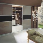 A luxurious walk-in wardrobe with sliding doors revealing organized shelves and hanging space, paired elegantly with a classical chandelier and an ornate bench.