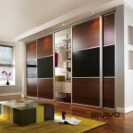 A modern sliding door wardrobe with mirrored and wooden panels in a tidy room featuring a sleek, wooden floor and a colorful cube-shaped ottoman. The wardrobe is open, displaying organized shelves and hanging spaces.