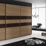 A modern bedroom interior featuring a large, segmented wooden wardrobe with brown stripes, complemented by bespoke furniture, a lush rug, and elegant chandelier lighting.