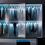 A modern, dimly lit walk-in closet featuring large fitted wardrobes with clear glass doors and internal lighting, showcasing an organized display of clothes and accessories.
