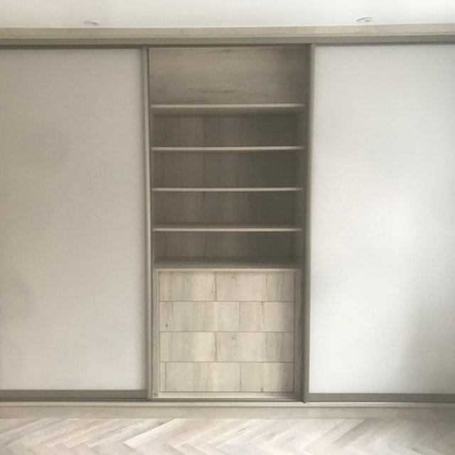 A sleek, minimalist Hampstead wardrobe with sliding doors. The doors are a light beige color, and the central compartment is open, revealing several shelves above two small drawer units. The flooring is herringbone patterned hardwood, complementing the wardrobe's design.