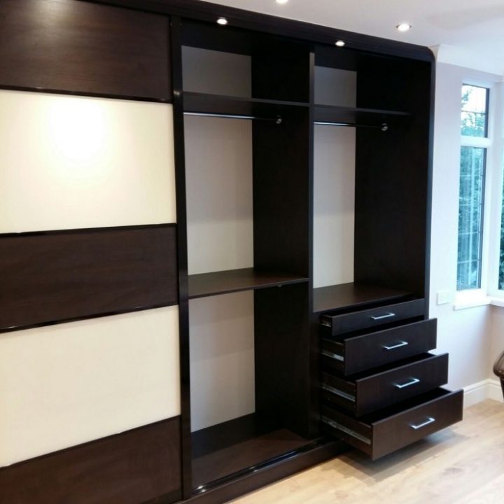 A modern, dark wooden fitted wardrobe with sliding doors, shelves, and built-in drawers, located in a room with light wood flooring and a bright lamp on the wall.