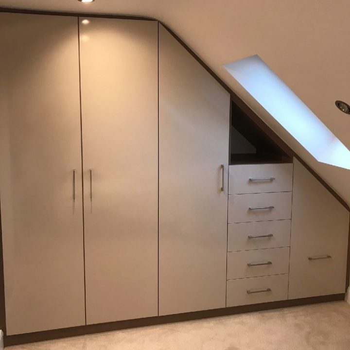 A custom-fitted wardrobe in Bravo style installed in an attic, featuring a slanted right side that aligns with the ceiling. The wardrobe has three vertical cabinet doors on the left and five horizontal drawers on the right, below a small skylight window on a beige carpeted floor in Walthamstow.