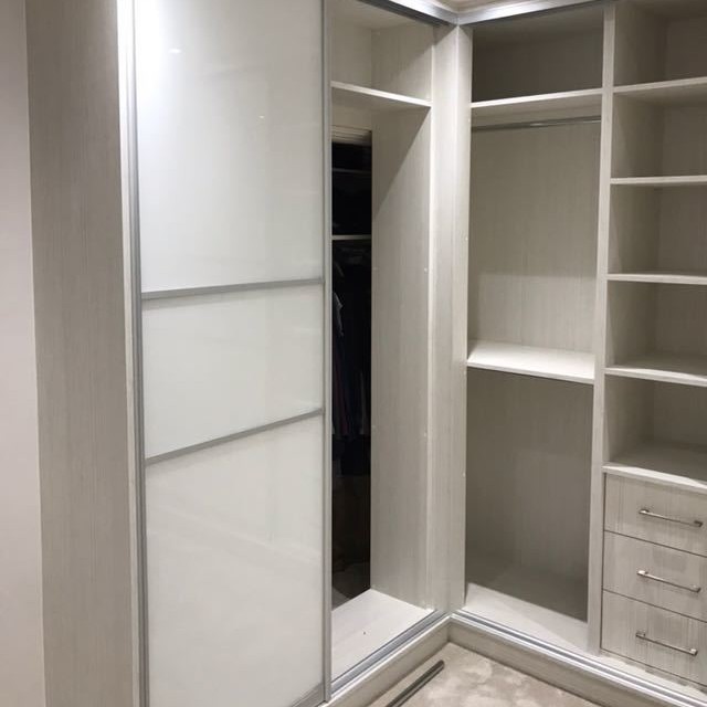 A modern walk-in closet with one hinged door partially open revealing clothes hanging inside. The closet features white shelves and drawers on the right.