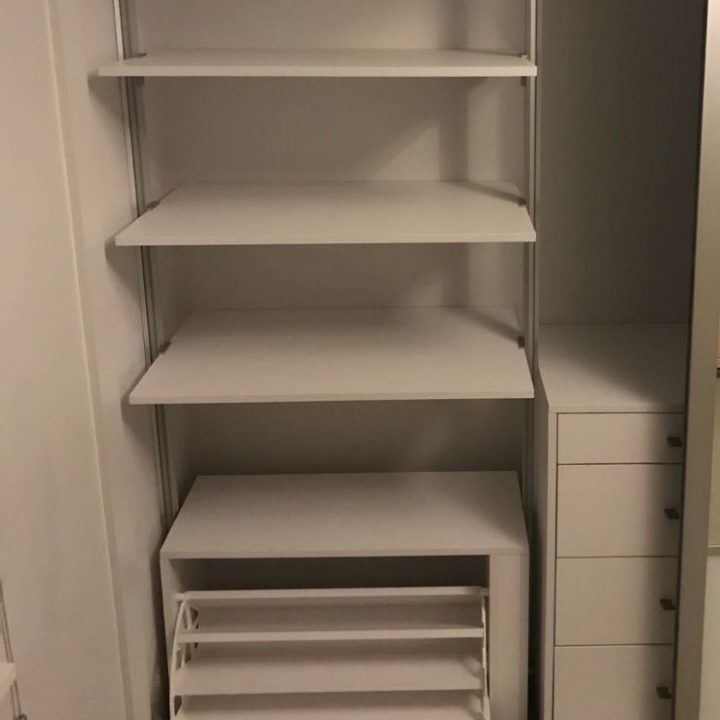 An empty white fitted wardrobe with multiple shelves and a built-in drawer unit, located in a room with dim lighting.