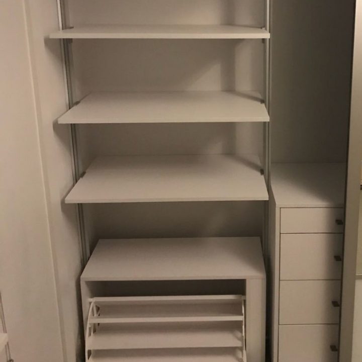 A built-in closet with white shelving units and sliding doors. The closet has several empty shelves and a four-drawer dresser on the right side. Below the shelves, there is an empty shoe rack. The overall look, complemented by the soft green hue of the doors, is clean and minimalistic.