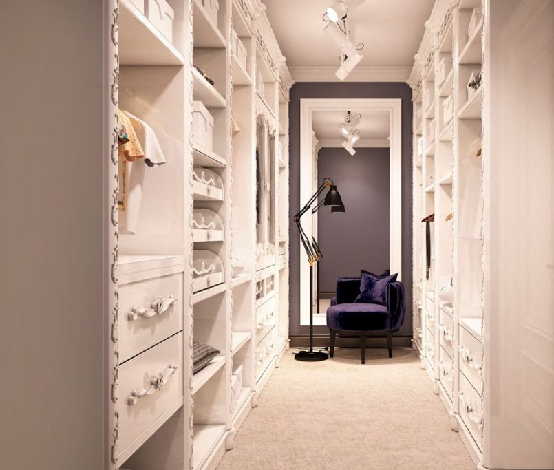 A spacious and well-lit walk-in closet with white built-in shelves and drawers filled with clothing and accessories on both sides. At the end of the closet is a seating area featuring a dark blue armchair and a floor lamp, set against a dark accent wall with a large mirror—a perfect blend of built in wardrobe vs walk in closet features.