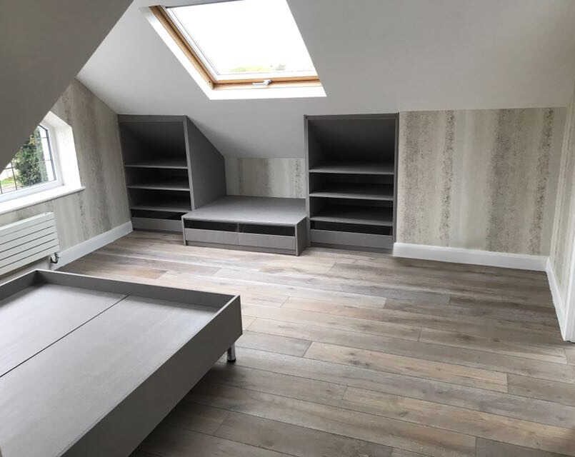 A modern, minimalist attic bedroom with grey storage units, light wood flooring, and a skylight. The room has built-in shelves and drawers, a single bed frame, and walls lined with subtle, textured wallpaper. Natural light illuminates the space—perfect for those seeking minimalist bedroom ideas.