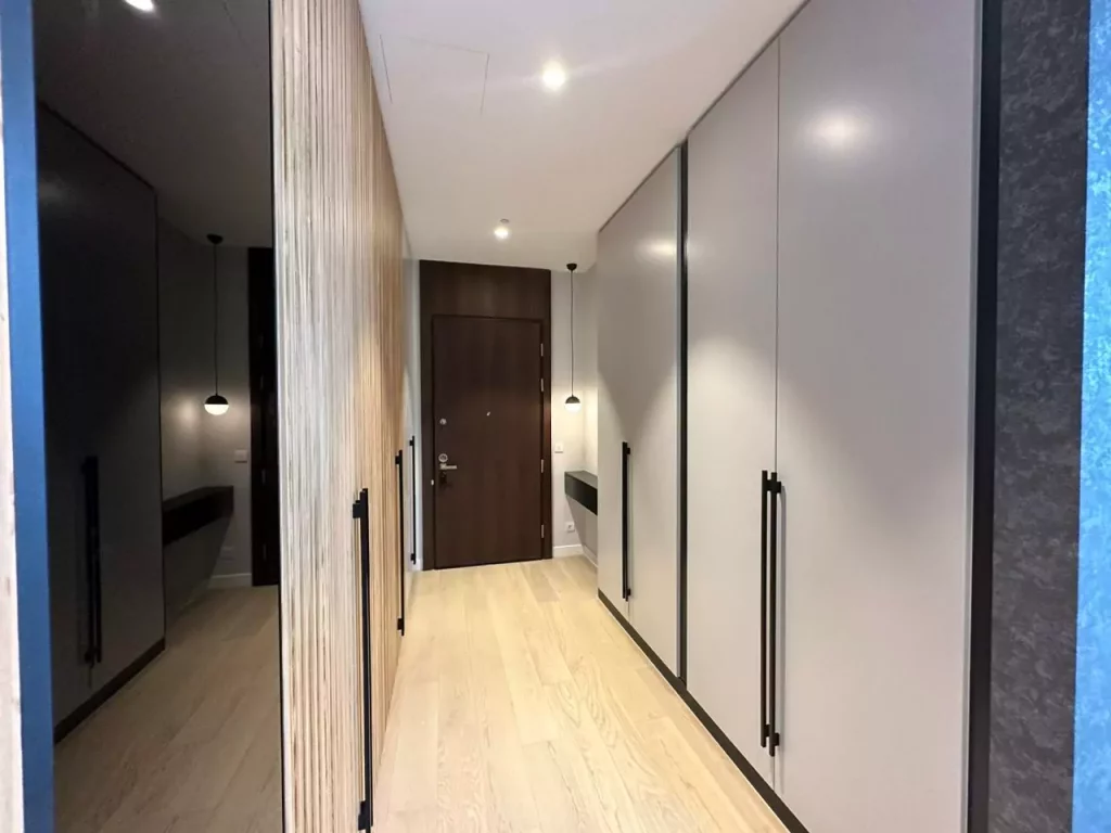 A sleek, modern hallway with light wood flooring and walls featuring vertical wooden slats on one side and smooth, grey cabinets with black handles on the other. At the end of the hallway is a dark wooden door, illuminated by a small wall-mounted light.