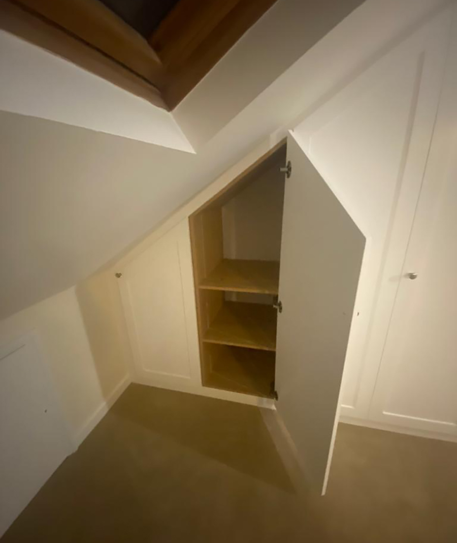 Your unique built-in wardrobe features a white closet with an open door revealing two wooden shelves inside. Built beneath a slanted ceiling, likely in an attic or upper floor, it is surrounded by white walls and a beige carpet. A skylight is visible at the top left corner, adding natural light.