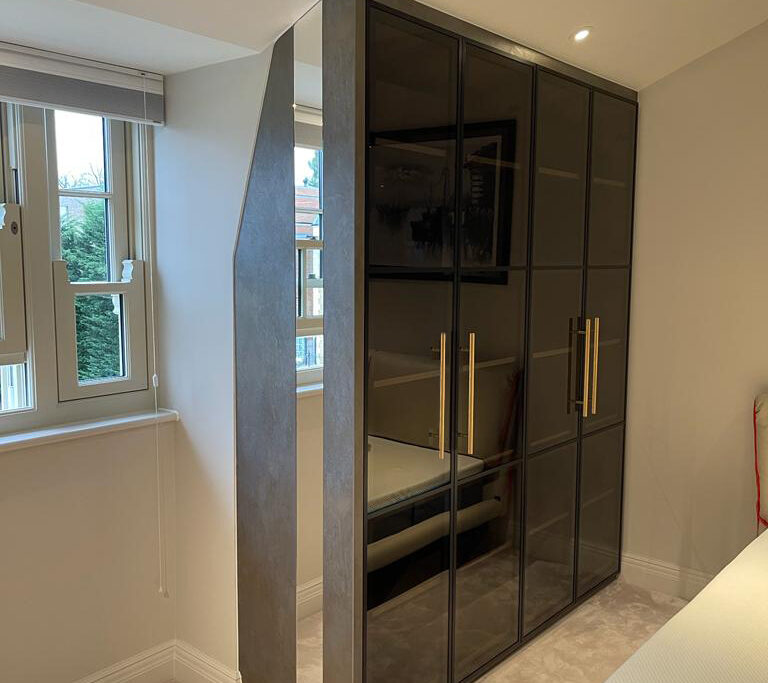 A bedroom showcasing exquisite interior design features a large wardrobe with reflective black glass doors and gold handles. Positioned against a wall near a window, the wardrobe complements a bed with a gray mattress and green pillow. Natural light streams in, illuminating the stylish room.