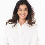 A woman with long, dark hair is smiling at the camera. She is wearing a white blouse and gold hoop earrings, embodying a casual, open and friendly demeanor. The background is plain white, reminiscent of the sleek design found in fitted bedrooms across London.
