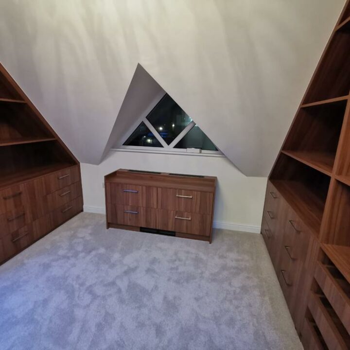 A cozy attic room with gray carpet featuring an angular window between two wooden, bespoke shelving units and a matching cabinet below the window.