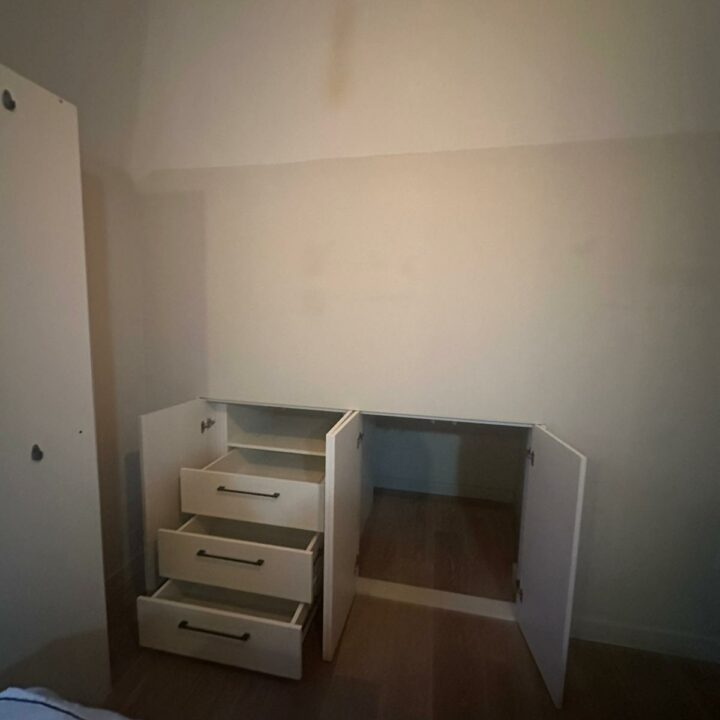 A dimly lit room featuring a white, partially dismantled dresser with three drawers lying on the floor. The walls show some discoloration and a fitted wardrobe is visible in the corner.