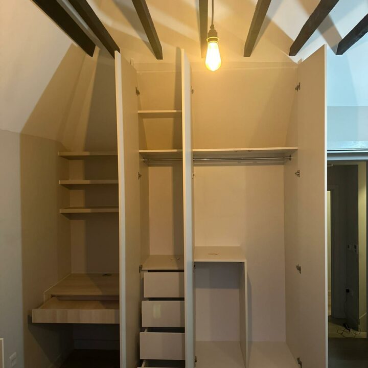 A minimalistic built-in closet with exposed beams, featuring shelves, drawers, a hanging rod, and an integrated ladder under a lit bulb has been transformed into a walk-in wardrobe.