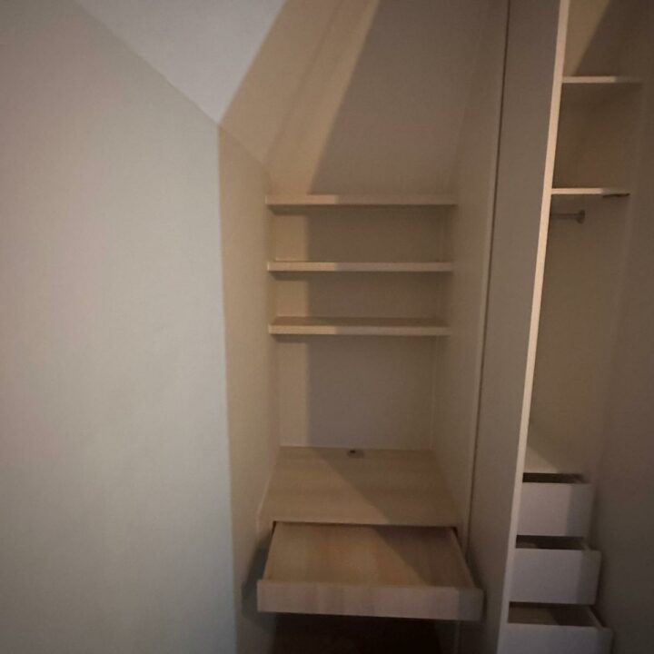 An under-staircase storage solution featuring bespoke wooden shelves on the left side with additional shelving seen ascending with the stairs on the right in a dimly lit environment.