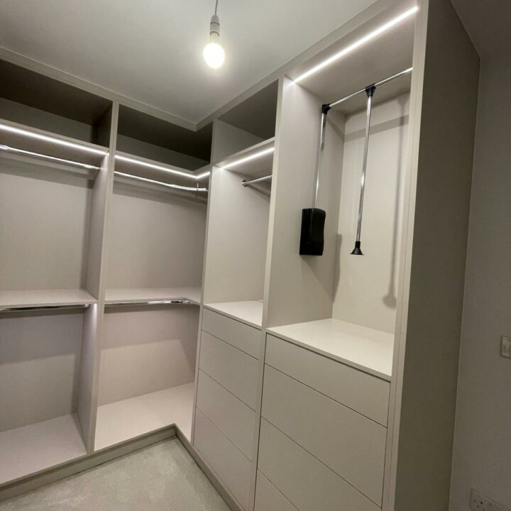 A modern walk-in wardrobe featuring empty shelves, drawers, and a hanging space with LED strip lighting, set in a room with gray walls and a textured gray floor.