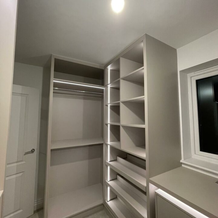 A modern, empty walk-in wardrobe featuring built-in shelves and hanging spaces, with a single overhead light bulb illuminating the area. The walls and cabinetry are in neutral tones.