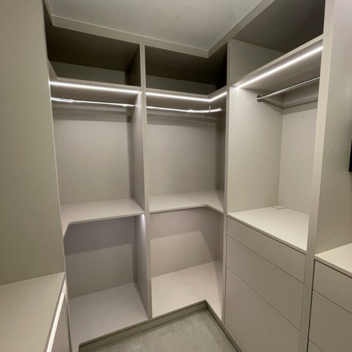 Empty walk-in closet with built-in white shelving units and hanging rods illuminated by overhead lighting, featuring bespoke furniture.