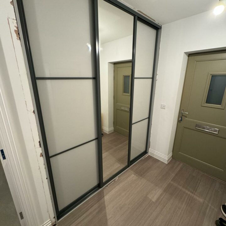 A modern interior featuring a large black-framed sliding door with frosted glass panels. The door is set against a backdrop of light gray walls and wooden flooring, complemented by bespoke furniture, with a green door visible in the background.