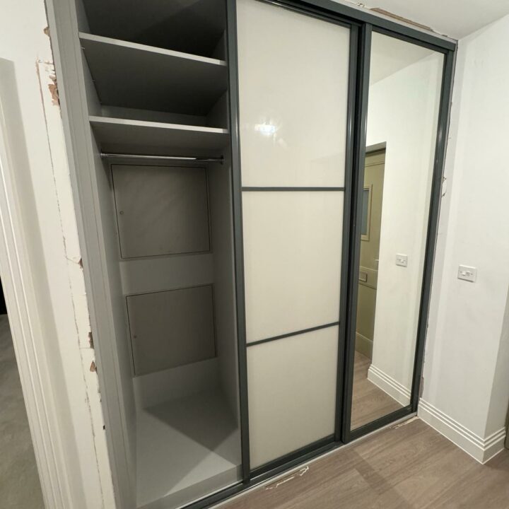 An empty built-in wardrobe with a large mirrored sliding door, partially open to reveal interior shelves and compartments, set in a room with laminate flooring and white walls. This is an essential component of our bespoke furniture collection for fitted bedrooms.